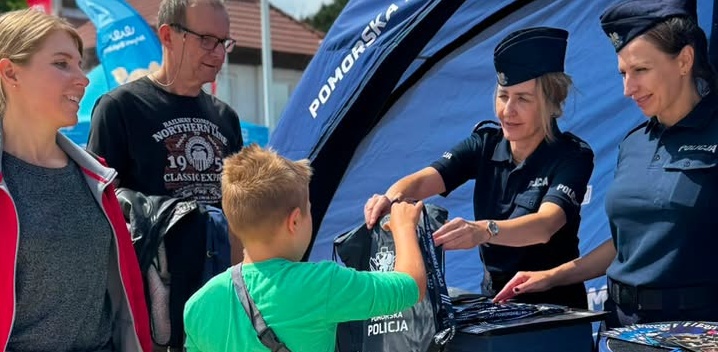 Policjantka w ciemnoniebieskim mundurze i furażerce wręcza czarny worek z logo chłopcu w zielonej koszulce przy stoisku profilaktycznym w Gdańsku-Brzeźnie w czasie akcji "Kręci mnie bezpieczeństwo w lecie". Obok stoją uśmiechnięci opiekunowie dziecka, w tle widać fragment niebieskiego namiotu policji