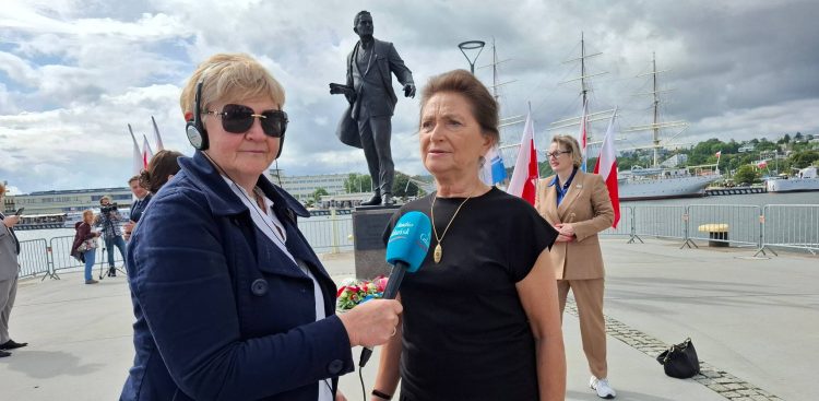 Dziennikarka Radia Gdańsk Anna Rębas w okularach przeciwsłonecznych i słuchawkach trzyma mikrofon z logo Radia Gdańsk, przeprowadzając wywiad z kobietą w czarnej bluzce i ze złotym wisiorkiem. To Hanna Wenda-Uszyńska, wnuczką Tadeusza Wendy. W tle, na placu przy nabrzeżu, widoczny jest pomnik Tadeusza Wendy, polskie flagi oraz cumujące w porcie żaglowce pod zachmurzonym niebem. To urodziny budowniczego gdyńskiego portu