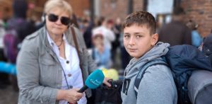 Młody chłopiec w szarej bluzie z plecakiem na ramionach patrzy w stronę obiektywu (uczestnik pielgrzymki z Gdańska na Jasną Górę). Obok niego stoi kobieta w okularach przeciwsłonecznych, trzymająca w ręku niebieski mikrofon z logo „Radio Gdańsk” (Anna Rębas). W rozmytym tle widać ceglany budynek oraz zgromadzony tłum ludzi z plecakami