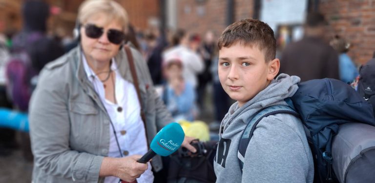 Młody chłopiec w szarej bluzie z plecakiem na ramionach patrzy w stronę obiektywu (uczestnik pielgrzymki z Gdańska na Jasną Górę). Obok niego stoi kobieta w okularach przeciwsłonecznych, trzymająca w ręku niebieski mikrofon z logo „Radio Gdańsk” (Anna Rębas). W rozmytym tle widać ceglany budynek oraz zgromadzony tłum ludzi z plecakami