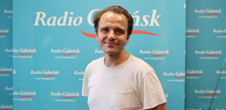 Uśmiechnięty mężczyzna z krótkim zarostem, ubrany w biały t-shirt z kieszonką, pozujący na tle turkusowej ścianki z logo Radio Gdańsk. Aktor Paweł Palcat