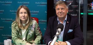 Kobieta w zielonej, wzorzystej marynarce (Beata Dunajewska) oraz mężczyzna w ciemnej marynarce i jasnej koszuli w paski (Piotr Karczewsk) siedzą przy stole w studiu radiowym przed mikrofonem. W tle widoczna jest niebieska ścianka z powtarzającym się logo "Radio Gdańsk"