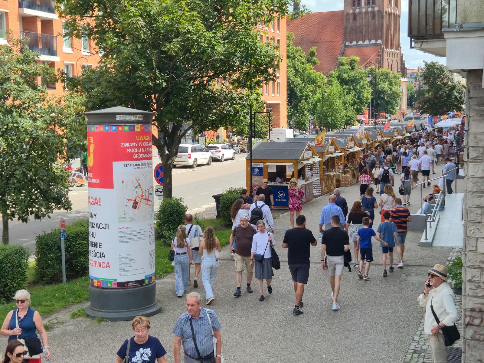 Jarmark św. Dominika w Gdańsku. Szeroki chodnik, wzdłuż którego ciągnie się rząd drewnianych domków jarmarcznych. Po deptaku spacerują ludzie w letnich ubraniach. Po lewej stronie stoi szary słup ogłoszeniowy z czerwonym plakatem informacyjnym, a w tle, nad zielonymi drzewami, wznosi się gotycka wieża kościoła z czerwonej cegły