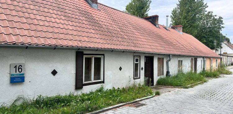 Checz rybacka w gdańskim Jelitkowie przed remontem. Parterowa zabytkowa chata rybacka o białych tynkowanych ścianach i spadzistym dachu z czerwoną dachówką. Na elewacji po lewej stronie wisi niebieska tabliczka adresowa z numerem 16 przy ulicy Bałtyckiej. Wzdłuż budynku i brukowanej drogi rośnie wysoka trawa i chwasty