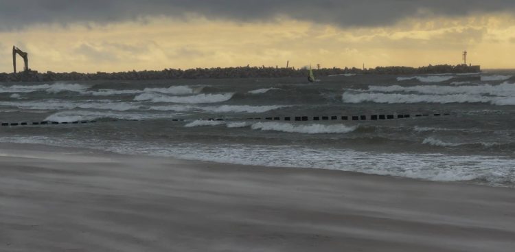 Sztormowe, spienione fale Bałtyku rozbijające się o plażę w Ustce. Na pierwszym planie drewniane ostrogi i piasek unoszony silnym wiatrem. W tle kamienny falochron z widoczną koparką budowlaną pod ciemnym, burzowym niebem. To wieczór przed zapowiadanym uderzeniem cyklonu Bernadette