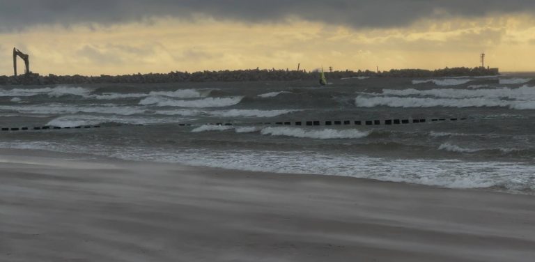 Sztormowe, spienione fale Bałtyku rozbijające się o plażę w Ustce. Na pierwszym planie drewniane ostrogi i piasek unoszony silnym wiatrem. W tle kamienny falochron z widoczną koparką budowlaną pod ciemnym, burzowym niebem. To wieczór przed zapowiadanym uderzeniem cyklonu Bernadette