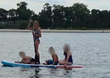 Czworo dzieci z zamazanymi twarzami na jednej desce SUP na wodzie. Jedno dziecko stoi i trzyma wiosło, pozostała trójka siedzi na desce. Żadne z dzieci nie ma na sobie kamizelki ratunkowej