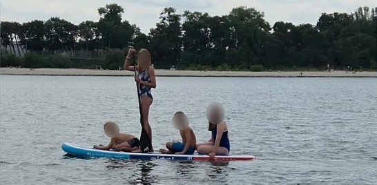 Czworo dzieci z zamazanymi twarzami na jednej desce SUP na wodzie. Jedno dziecko stoi i trzyma wiosło, pozostała trójka siedzi na desce. Żadne z dzieci nie ma na sobie kamizelki ratunkowej