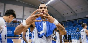Koszykarz Dominic Green w biało-niebieskim stroju MKS Dąbrowa Górnicza i z niebieskim ręcznikiem na szyi, wykonujący gest dłońmi w stronę obiektywu na tle hali sportowej. Zdjęcie ilustruje przejście zawodnika do klubu Energa Czarni Słupsk