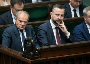 Fotografia przedstawia polityków zasiadających w ławach rządowych w sali plenarnej Sejmu. W pierwszym rzędzie widoczni są od lewej: premier Donald Tusk z poważnym, zatroskanym wyrazem twarzy, wicepremier Władysław Kosiniak-Kamysz opierający brodę na dłoni oraz minister spraw wewnętrznych i administracji Tomasz Siemoniak trzymający dłoń przy uchu. Wszyscy ubrani są w ciemne garnitury i krawaty. W tle, na zielonych fotelach sejmowych, siedzą inni członkowie rządu i urzędnicy