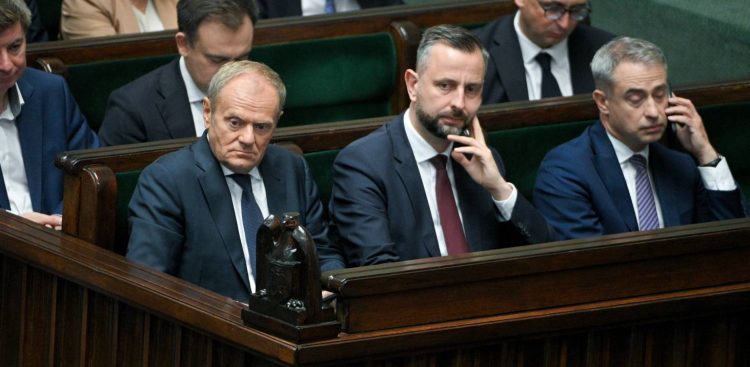 Fotografia przedstawia polityków zasiadających w ławach rządowych w sali plenarnej Sejmu. W pierwszym rzędzie widoczni są od lewej: premier Donald Tusk z poważnym, zatroskanym wyrazem twarzy, wicepremier Władysław Kosiniak-Kamysz opierający brodę na dłoni oraz minister spraw wewnętrznych i administracji Tomasz Siemoniak trzymający dłoń przy uchu. Wszyscy ubrani są w ciemne garnitury i krawaty. W tle, na zielonych fotelach sejmowych, siedzą inni członkowie rządu i urzędnicy