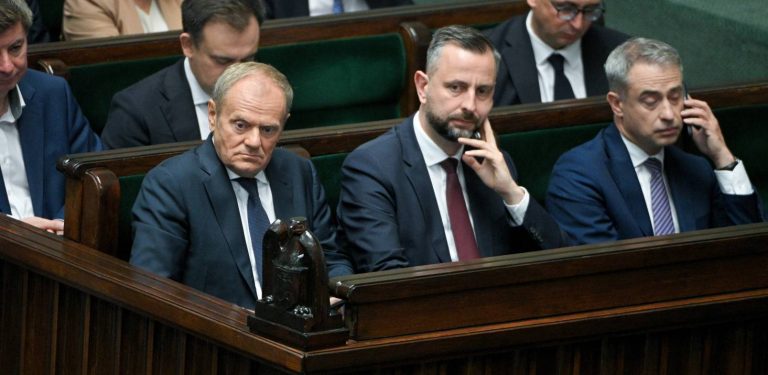Fotografia przedstawia polityków zasiadających w ławach rządowych w sali plenarnej Sejmu. W pierwszym rzędzie widoczni są od lewej: premier Donald Tusk z poważnym, zatroskanym wyrazem twarzy, wicepremier Władysław Kosiniak-Kamysz opierający brodę na dłoni oraz minister spraw wewnętrznych i administracji Tomasz Siemoniak trzymający dłoń przy uchu. Wszyscy ubrani są w ciemne garnitury i krawaty. W tle, na zielonych fotelach sejmowych, siedzą inni członkowie rządu i urzędnicy