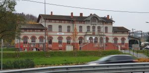 Zabytkowy, piętrowy budynek dworca kolejowego w Redzie z jasnoszarą elewacją, czerwonymi, ceglanymi zdobieniami wokół okien i u podstawy. Przed budynkiem ciągnie się zielony trawnik z młodymi drzewkami, a po prawej stronie widać schody wejściowe. Na pierwszym planie widoczny jest rozmyty w ruchu, jadący samochód. Dworzec w redzie ma znów być dostępny dla pasażerów