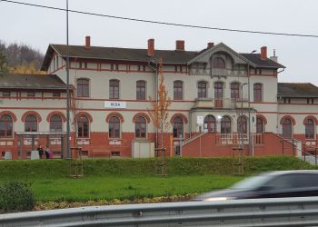 Zabytkowy, piętrowy budynek dworca kolejowego w Redzie z jasnoszarą elewacją, czerwonymi, ceglanymi zdobieniami wokół okien i u podstawy. Przed budynkiem ciągnie się zielony trawnik z młodymi drzewkami, a po prawej stronie widać schody wejściowe. Na pierwszym planie widoczny jest rozmyty w ruchu, jadący samochód. Dworzec w redzie ma znów być dostępny dla pasażerów