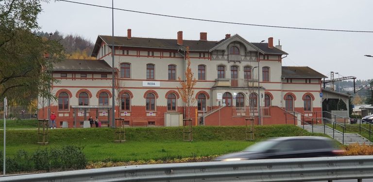 Zabytkowy, piętrowy budynek dworca kolejowego w Redzie z jasnoszarą elewacją, czerwonymi, ceglanymi zdobieniami wokół okien i u podstawy. Przed budynkiem ciągnie się zielony trawnik z młodymi drzewkami, a po prawej stronie widać schody wejściowe. Na pierwszym planie widoczny jest rozmyty w ruchu, jadący samochód. Dworzec w redzie ma znów być dostępny dla pasażerów