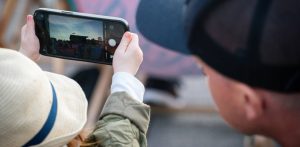 Dziecko w słomkowym kapeluszu trzyma oburącz smartfon, robiąc zdjęcie plenerowego koncertu o zmierzchu. Obok niego, tyłem do obiektywu, stoi osoba w ciemnej czapce z daszkiem.