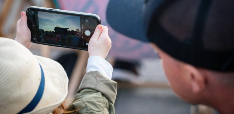 Dziecko w słomkowym kapeluszu trzyma oburącz smartfon, robiąc zdjęcie plenerowego koncertu o zmierzchu. Obok niego, tyłem do obiektywu, stoi osoba w ciemnej czapce z daszkiem.