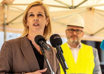 Emilia Lodzińska, zastępczyni prezydent Gdańska, przemawia do dziennikarzy na tle rozmytego wizerunku mężczyzny w okularach, białym kasku ochronnym i żółtej kamizelce odblaskowej. Spotkanie odbywa się pod beżowym zadaszeniem przy ul. Szczyglej 2 w Gdańsku, gdzie powstają nowe mieszkania TBS Motława