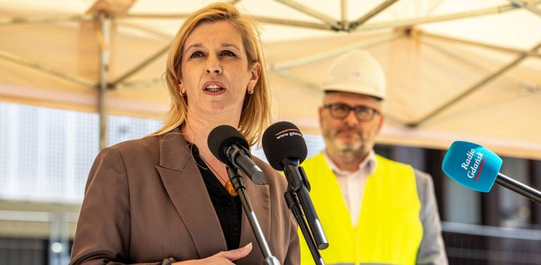 Emilia Lodzińska, zastępczyni prezydent Gdańska, przemawia do dziennikarzy na tle rozmytego wizerunku mężczyzny w okularach, białym kasku ochronnym i żółtej kamizelce odblaskowej. Spotkanie odbywa się pod beżowym zadaszeniem przy ul. Szczyglej 2 w Gdańsku, gdzie powstają nowe mieszkania TBS Motława