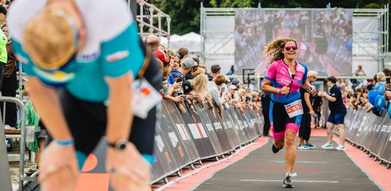 Kobieta w różowo-niebieskim stroju triathlonowym klubu TriToruń z numerem startowym 652 biegnie z uśmiechem po czerwonym dywanie w strefie mety Enea IRONMAN Gdynia 2023. Po bokach stoją kibice oparci o barierki. Na pierwszym planie, po lewej stronie, widać rozmytą, pochyloną sylwetkę innego, zmęczonego zawodnika. W tle widoczny jest duży ekran LED