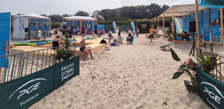 Farma Dobrej Energii na plaży w Ustce. Drewniane podesty, leżaki, pufy i zadaszone stoiska informacyjne Fundacji PGE w strefie „Farma Dobrej Energii” na plaży w Ustce, wokół której wypoczywają dorośli i dzieci