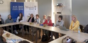 Grupa ludzi siedzi przy stołach podczas spotkania w jasnej sali. W tle stoją dwa roll-upy: niebieski z logo „Gdańsk Wybiera Zdrowie” oraz biały z napisem „Wrzeszcz Dolny Rada Dzielnicy”. Po prawej stronie przy laptopie siedzi prelegentka w żółtej żakiecie