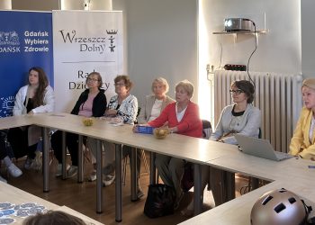 Grupa ludzi siedzi przy stołach podczas spotkania w jasnej sali. W tle stoją dwa roll-upy: niebieski z logo „Gdańsk Wybiera Zdrowie” oraz biały z napisem „Wrzeszcz Dolny Rada Dzielnicy”. Po prawej stronie przy laptopie siedzi prelegentka w żółtej żakiecie