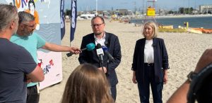Mariusz Bzdęga i Jagna Ogonowska podczas konferencji prasowej na plaży w Gdyni. Pośrodku stoi mężczyzna w okularach i ciemnej marynarce, mówiący do kilku trzymanych przez dziennikarzy mikrofonów. Po jego prawej stronie stoi uśmiechnięta blondynka w ciemnym żakiecie i białej bluzce. W tle widać plażę, morze, żółtą wieżę ratowniczą oraz fragmenty banerów reklamowych
