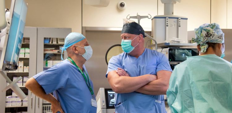 Dwóch lekarzy w jasnoniebieskich strojach chirurgicznych, maseczkach ochronnych i czepkach rozmawia ze sobą w sali szpitalnej. Lekarz po lewej, starszy mężczyzna, ma na szyi zieloną smycz z wyraźnym białym napisem „LEKARZ” oraz przypięty identyfikator. Lekarz po prawej stoi ze skrzyżowanymi ramionami i słucha rozmówcy. Po prawej stronie kadru tyłem do obiektywu stoi trzecia postać w zielonym fartuchu medycznym i wzorzystym czepku. W tle widoczne są elementy wyposażenia szpitalnego: ekrany diagnostyczne, szafki z materiałami medycznymi oraz aparatura zabiegowa. To Wojewódzki Szpital Specjalistyczny im. Janusza Korczaka w Słupsku Sp. z o.o., oddział ginekologiczny