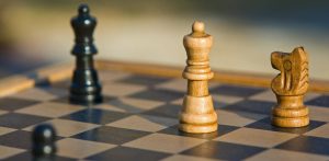 Zbliżenie na drewnianą szachownicę w ciepłym, naturalnym świetle. W ostrości po prawej stronie stoją dwie jasne figury: hetman (królowa) oraz skoczek (koń). W tle, po lewej stronie, widoczna jest rozmyta czarna figura, a w lewym dolnym rogu kadru lekko zarysowany czarny pionek