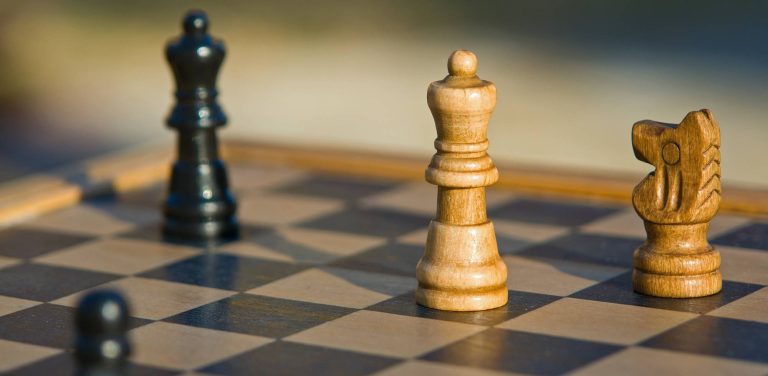 Zbliżenie na drewnianą szachownicę w ciepłym, naturalnym świetle. W ostrości po prawej stronie stoją dwie jasne figury: hetman (królowa) oraz skoczek (koń). W tle, po lewej stronie, widoczna jest rozmyta czarna figura, a w lewym dolnym rogu kadru lekko zarysowany czarny pionek