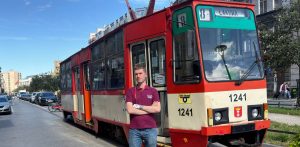 Młody mężczyzna ze skrzyżowanymi rękami, ubrany w bordową koszulkę polo z logo i jeansy, stoi przed zabytkowym, czerwono-kremowym tramwajem o numerze bocznym 1241. Na tramwaju widnieje tablica linii 8 z kierunkiem "Łąkowa", logo ZKM Gdańsk oraz herb Gdańska. W tle widać miejską ulicę, zaparkowane samochody i kamienice. Mężczyzna to Cezary Kamiński, prezes stowarzyszenia Grupa Tramwajowo-Autobusowa WPK GG, którego celem jest utworzenie Muzeum Komunikacji w Gdańsku