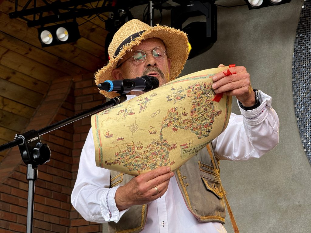 Mężczyzna w kapeluszu, uczestnik Mistrzostw w Zażywaniu Tabaki, przmawia do mikrofonu, w rękach trzymając mapę
