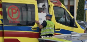 Zbliżenie na żółto-czerwoną karetkę pogotowia ratunkowego z otwartymi bocznymi drzwiami. Przy pojeździe stoi policjant w białej czapce i jaskrawozielonej kamizelce odblaskowej z napisem „POLICJA”, zaglądający do wnętrza ambulansu. Na szybie pojazdu medycznego widoczne jest czerwone oznaczenie z literą „P” oraz numer G 0204. Informacja o włamywaczu, który wyskoczył z okna plebanii w powiecie słupskim