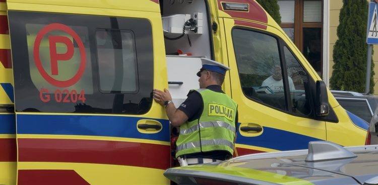 Zbliżenie na żółto-czerwoną karetkę pogotowia ratunkowego z otwartymi bocznymi drzwiami. Przy pojeździe stoi policjant w białej czapce i jaskrawozielonej kamizelce odblaskowej z napisem „POLICJA”, zaglądający do wnętrza ambulansu. Na szybie pojazdu medycznego widoczne jest czerwone oznaczenie z literą „P” oraz numer G 0204. Informacja o włamywaczu, który wyskoczył z okna plebanii w powiecie słupskim
