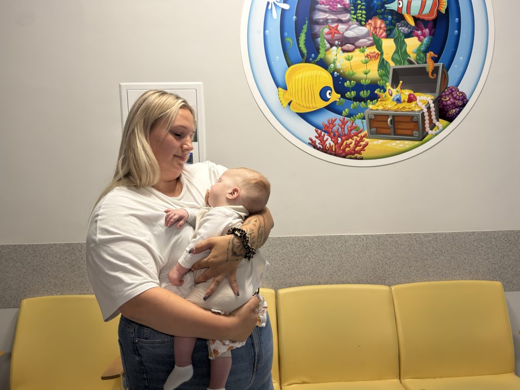 Kobieta trzymająca na rękach niemowlę w Uniwersyteckim Centrum Klinicznym, w tle widoczne żółte fotele i kolorowa ścienna grafika z motywem podwodnym.