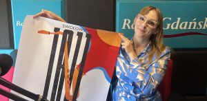 Uśmiechnięta kobieta w okularach i niebiesko-białej bluzce prezentuje i wskazuje plakat festiwalu "Swołowo Jazz (to Gabriela Kalinowska z Centrum Kultury i Biblioteki Gminy Redzikowo). Kobieta znajduje się w studiu radiowym, w tle widać logo Radia Gdańsk, a z lewej strony widoczny jest mikrofon