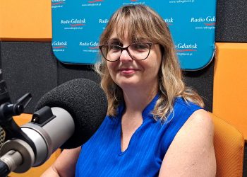 Dr Justyna Kobos w studiu Radia Gdańsk. Kobieta ma półdługie, jasne włosy z grzywką, okulary w ciemnych oprawkach i niebieską bluzkę bez rękawów. Patrzy w stronę obiektywu z lekkim uśmiechem. Na pierwszym planie po lewej stronie widoczny jest profesjonalny mikrofon studyjny z czarną gąbką. W tle znajdują się pomarańczowe i czarne panele akustyczne oraz niebieska tablica z napisami „Radio Gdańsk”. Rozmowa dotyczy sinic, które mogą być niebezpieczne dla turystów kąpiących się w morzu
