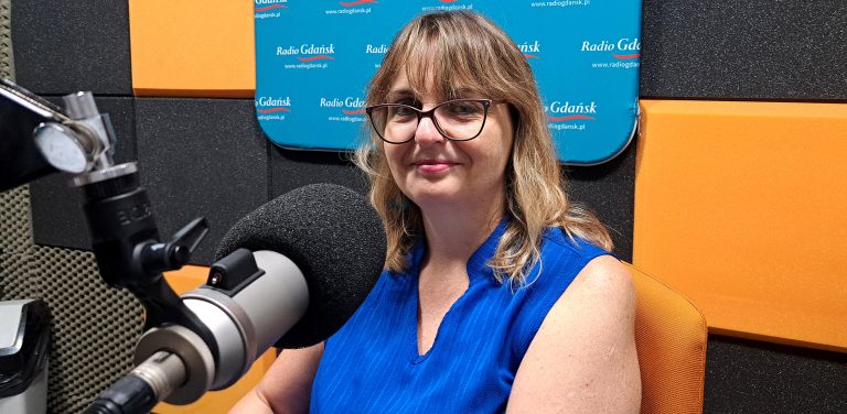 Dr Justyna Kobos w studiu Radia Gdańsk. Kobieta ma półdługie, jasne włosy z grzywką, okulary w ciemnych oprawkach i niebieską bluzkę bez rękawów. Patrzy w stronę obiektywu z lekkim uśmiechem. Na pierwszym planie po lewej stronie widoczny jest profesjonalny mikrofon studyjny z czarną gąbką. W tle znajdują się pomarańczowe i czarne panele akustyczne oraz niebieska tablica z napisami „Radio Gdańsk”. Rozmowa dotyczy sinic, które mogą być niebezpieczne dla turystów kąpiących się w morzu
