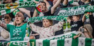 Zbliżenie na młodą dziewczynkę w jasnej bluzie w różowe serduszka, stojącą na trybunach stadionu i trzymającą oburącz nad głową zielono-biały szalik z napisem „MŁODA LECHIA”. Wokół niej widoczni są inni kibice w różnym wieku, również trzymający w górze klubowe szaliki i dopingujący drużynę