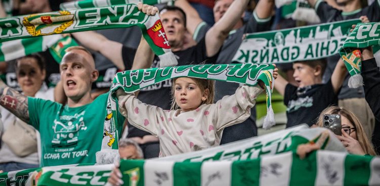 Zbliżenie na młodą dziewczynkę w jasnej bluzie w różowe serduszka, stojącą na trybunach stadionu i trzymającą oburącz nad głową zielono-biały szalik z napisem „MŁODA LECHIA”. Wokół niej widoczni są inni kibice w różnym wieku, również trzymający w górze klubowe szaliki i dopingujący drużynę
