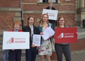 Grupa aktywistów stowarzyszenia Społeczny Gdańsk oraz partii Razem stojąca przed ceglanym budynkiem Rady Miasta Gdańska. Trzymają w dłoniach dokumenty projektu uchwały oraz białe i czerwone tabliczki z napisem „Społeczny GDAŃSK”. Celem inicjatywy jest zmiana obowiązującego Regulaminu Obywatelskich Inicjatyw Uchwałodawczych, uchwalonego w 2019 roku