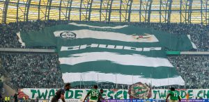Wielka sektorówka kibiców Lechii Gdańsk w kształcie klubowej, zielono-białej koszulki z napisem „CHWDP” rozpostarta na trybunie stadionu podczas meczu piłkarskiego. Na dole widoczny jest transparent z hasłem „TWARDE PIEŚCI...”, a na pierwszym planie zawodnicy biegający po murawie