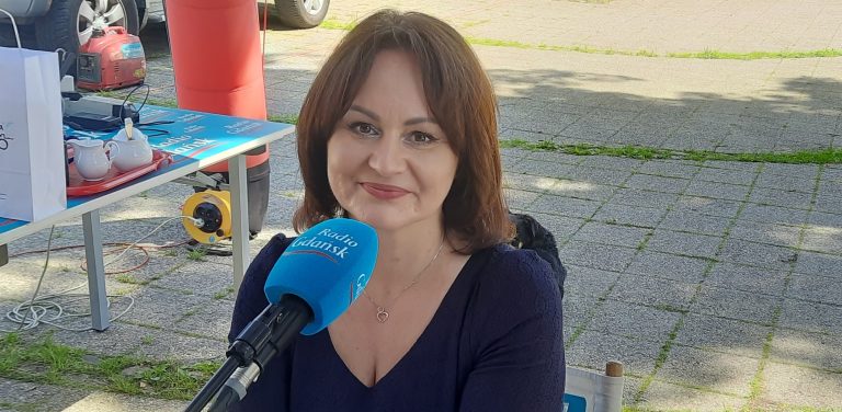 Uśmiechnięta kobieta z brązowymi włosami do ramion, w ciemnej bluzce z dekoltem V, siedząca przed mikrofonem z niebieską gąbką z logo Radia Gdańsk (burmistrz Żukowa Mariola Zmudzińska . W tle widać plenerowe studio radiowe na brukowanym placu