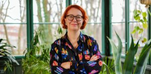 Portret uśmiechniętej kobiety w czarnych, okrągłych okularach i z rudymi włosami, stojącej z założonymi rękami na tle jasnego, przeszklonego pomieszczenia z roślinami. Kobieta ma na sobie ciemną koszulę w wyraziste, kolorowe wzory geometryczne. To Marta Czarnecka, nowa dyrektorka Goyki 3 Art Inkubatora w Sopocie