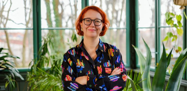 Portret uśmiechniętej kobiety w czarnych, okrągłych okularach i z rudymi włosami, stojącej z założonymi rękami na tle jasnego, przeszklonego pomieszczenia z roślinami. Kobieta ma na sobie ciemną koszulę w wyraziste, kolorowe wzory geometryczne. To Marta Czarnecka, nowa dyrektorka Goyki 3 Art Inkubatora w Sopocie