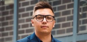 Portret młodego mężczyzny w ciemnej marynarce i jasnej koszuli, który patrzy w stronę obiektywu i lekko się uśmiecha. Na zdjęciu znajduje się Mateusz Klebba - tłumacz przysięgły języka kaszubskiego