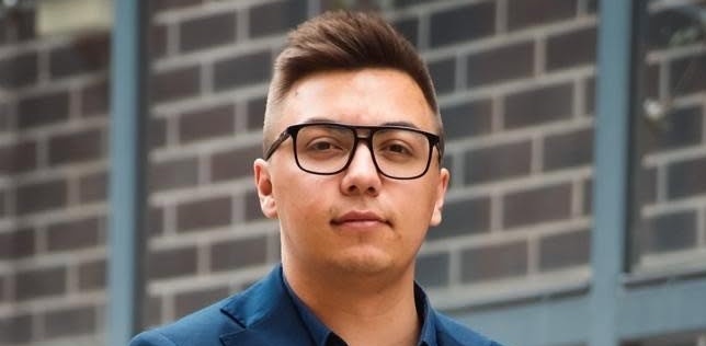 Portret młodego mężczyzny w ciemnej marynarce i jasnej koszuli, który patrzy w stronę obiektywu i lekko się uśmiecha. Na zdjęciu znajduje się Mateusz Klebba - tłumacz przysięgły języka kaszubskiego