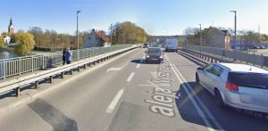 Widok z perspektywy jezdni na most Kardynała Wyszyńskiego w Elblągu. Na asfalcie widnieje namalowany napis „aleja Wyszyńskiego”. Po drodze poruszają się samochody, a po lewej stronie, na wydzielonym barierami chodniku, stoją dwie osoby. W tle po lewej widać rzekę, drzewa oraz wysoką wieżę kościoła