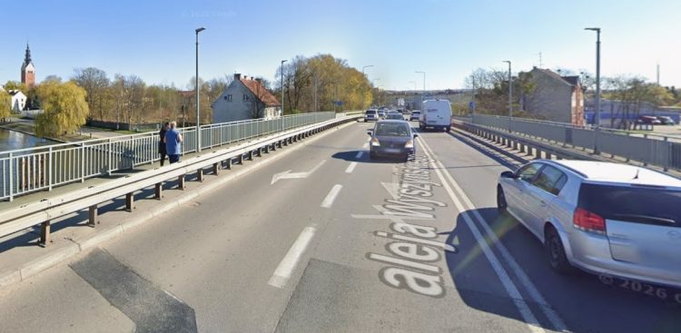 Widok z perspektywy jezdni na most Kardynała Wyszyńskiego w Elblągu. Na asfalcie widnieje namalowany napis „aleja Wyszyńskiego”. Po drodze poruszają się samochody, a po lewej stronie, na wydzielonym barierami chodniku, stoją dwie osoby. W tle po lewej widać rzekę, drzewa oraz wysoką wieżę kościoła